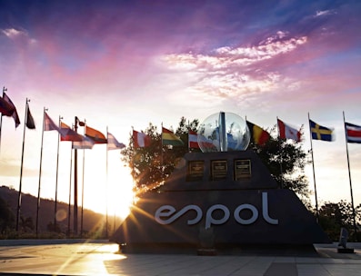 A sculpture with the logo 'ESPOL' is surrounded by numerous international flags. The sun is setting behind the sculpture, casting warm light and shadows across the scene. A clear sky with some clouds is visible in the background.