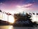 A sculpture with the logo 'ESPOL' is surrounded by numerous international flags. The sun is setting behind the sculpture, casting warm light and shadows across the scene. A clear sky with some clouds is visible in the background.