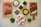 An assortment of fresh ingredients is neatly arranged on a light-colored kitchen surface. On the left, a wooden board displays slices of pepperoni and ham. In the center, small bowls contain grated cheese, spinach leaves, salsa, cooked minced meat, and corn kernels. To the right, a second wooden board holds sliced black olives, tomatoes, green peppers, and onions. A bowl of eggs is also part of the arrangement, accompanied by small piles of chopped green onions and herbs.