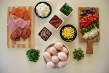 An assortment of fresh ingredients is neatly arranged on a light-colored kitchen surface. On the left, a wooden board displays slices of pepperoni and ham. In the center, small bowls contain grated cheese, spinach leaves, salsa, cooked minced meat, and corn kernels. To the right, a second wooden board holds sliced black olives, tomatoes, green peppers, and onions. A bowl of eggs is also part of the arrangement, accompanied by small piles of chopped green onions and herbs.