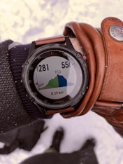 A person's wrist wearing a black Garmin watch displaying an elevation map of a hiking trail. The screen shows elevation data in meters, with two numbers 1281 and 551. The distance covered is 8.59 km. The person is also wearing a brown leather glove and a dark fabric sleeve. Snow is visible in the background.