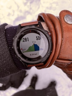 A person's wrist wearing a black Garmin watch displaying an elevation map of a hiking trail. The screen shows elevation data in meters, with two numbers 1281 and 551. The distance covered is 8.59 km. The person is also wearing a brown leather glove and a dark fabric sleeve. Snow is visible in the background.
