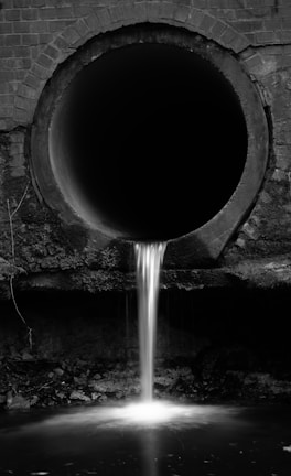 A black and white image featuring a large, circular drainage pipe embedded in a brick wall. Water cascades steadily from the pipe, flowing down into a pool below. The surrounding area appears damp and slightly overgrown, with some visible moss or algae.