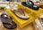 Three pairs of sandals are displayed on a glass surface with yellow boxes. The sandals have different brown and black straps and soles. There are small decorative lights adorning the boxes. In the background, various other footwear is visible on shelves.