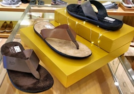 Three pairs of sandals are displayed on a glass surface with yellow boxes. The sandals have different brown and black straps and soles. There are small decorative lights adorning the boxes. In the background, various other footwear is visible on shelves.
