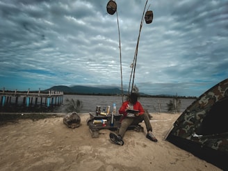 A person in a red jacket is sitting on sandy ground near a tent and a wooden pier extending into the water. They are surrounded by camping gear and fishing rods. The sky is overcast, and mountains are visible in the background.