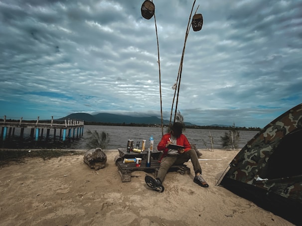 A person in a red jacket is sitting on sandy ground near a tent and a wooden pier extending into the water. They are surrounded by camping gear and fishing rods. The sky is overcast, and mountains are visible in the background.
