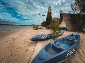 Two blue kayaks are resting on a sandy beach next to small huts with thatched roofs and green vegetation. The scene is set near a calm body of water under a cloudy sky.