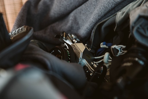 A vintage camera partially tucked inside a padded bag alongside various items. The scene includes soft fabrics and the camera lens, providing a glimpse of the brand and settings on the lens. The overall setting suggests preparation for travel or photography.