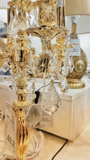 A close-up view of an intricate, gold-colored lamp adorned with crystal ornaments. The lamp is highly decorative, featuring elaborate carvings and designs. In the background, other lamps are visible, with price tags showing in the foreground.