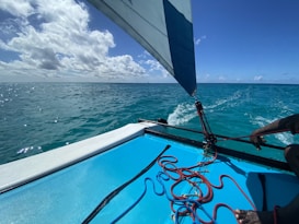A vibrant seascape featuring a sailboat with visible rigging and ropes. The boat's deck is painted in light blue, contrasting with the deep blue ocean. The sky is mostly clear with scattered clouds, and the sunlight creates sparkling reflections on the water's surface.