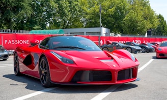 A sleek red sports car is prominently positioned in a parking lot filled with other high-end vehicles. Surrounding the area are banners and fences, and lush trees are visible in the background under a clear blue sky.