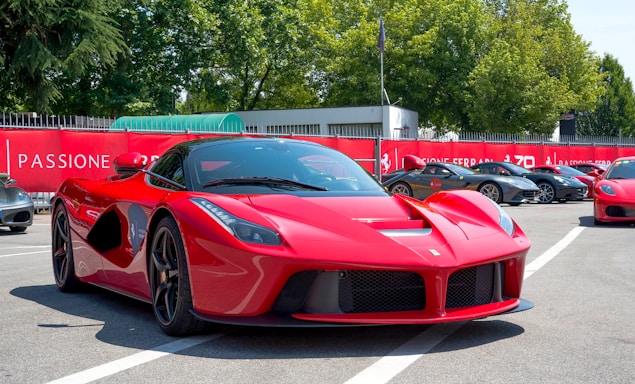 A sleek red sports car is prominently positioned in a parking lot filled with other high-end vehicles. Surrounding the area are banners and fences, and lush trees are visible in the background under a clear blue sky.