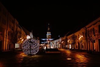 A festive urban scene at night featuring a large spherical Christmas decoration adorned with colorful lights in the foreground. The street is lined with bare trees and classical buildings, illuminated by warm street lamps. In the background, a tower is visible, decorated with additional festive lights.