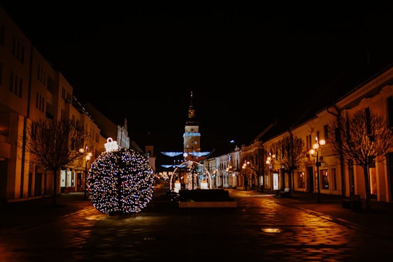 A festive urban scene at night featuring a large spherical Christmas decoration adorned with colorful lights in the foreground. The street is lined with bare trees and classical buildings, illuminated by warm street lamps. In the background, a tower is visible, decorated with additional festive lights.