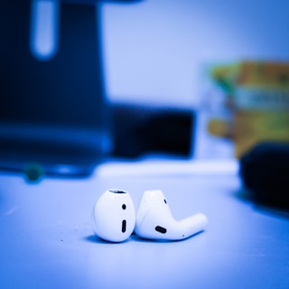 A pair of wireless earbuds lying on a flat surface in a room with blurred background elements. The focus is primarily on the earbuds, which are white and sleek, with a modern design. The lighting casts a cool blue tone over the entire scene.