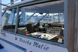 A close-up of a boat's cabin, with the name 'Hoocha Matic' visible on the side. The interior shows a cluttered dashboard with various equipment and tools. The steering wheel is noticeable, and the cabin window reflects the marina and boats in the background.