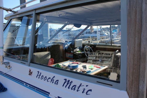 A close-up of a boat's cabin, with the name 'Hoocha Matic' visible on the side. The interior shows a cluttered dashboard with various equipment and tools. The steering wheel is noticeable, and the cabin window reflects the marina and boats in the background.