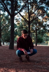 man in red hoodie and blue denim jeans sitting on brown ground near green trees during