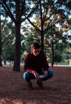 man in red hoodie and blue denim jeans sitting on brown ground near green trees during