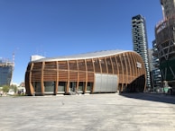 An architectural structure with a curved, wooden facade is set against a clear blue sky. The modern building is surrounded by other high-rise buildings under construction, with cranes visible in the background. There is a large open plaza area in front of the building, partially shaded.