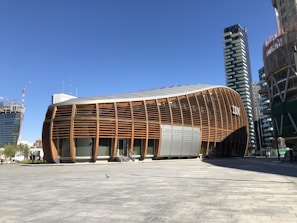 An architectural structure with a curved, wooden facade is set against a clear blue sky. The modern building is surrounded by other high-rise buildings under construction, with cranes visible in the background. There is a large open plaza area in front of the building, partially shaded.