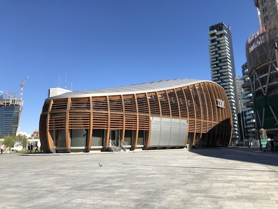 An architectural structure with a curved, wooden facade is set against a clear blue sky. The modern building is surrounded by other high-rise buildings under construction, with cranes visible in the background. There is a large open plaza area in front of the building, partially shaded.