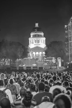 A monochrome photograph features a large crowd of people gathered in front of a stage at an outdoor event. The illuminated dome of a classical building serves as a backdrop, with trees partially obscuring its base. Stage equipment, including speakers and musical instruments, is visible, and stage lighting adds dramatic contrast.