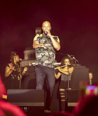 A performer stands on stage holding a microphone, dressed in a camouflage shirt and dark pants. Two women are visible in the background, one with braided hair and colorful attire, and the other playing a flute. The scene is set against a dark backdrop with stage lighting.