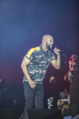 A performer dressed in a camouflage-patterned shirt sings passionately into a microphone on stage, while a cameraman records the event. The background is dark, emphasizing the spotlight on the performer.