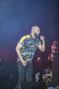 A performer dressed in a camouflage-patterned shirt sings passionately into a microphone on stage, while a cameraman records the event. The background is dark, emphasizing the spotlight on the performer.