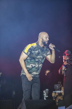 A performer dressed in a camouflage-patterned shirt sings passionately into a microphone on stage, while a cameraman records the event. The background is dark, emphasizing the spotlight on the performer.