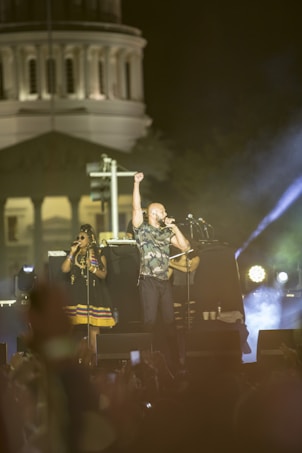 A musical performance taking place outdoors at night, featuring a male singer in camouflage holding his fist in the air and singing into a microphone. A female singer stands beside him, wearing sunglasses and a colorful dress. Bright stage lights and a large building in the background create an atmospheric setting, with a crowd visible in the foreground.