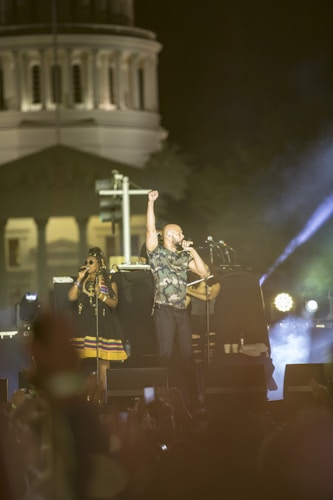 A musical performance taking place outdoors at night, featuring a male singer in camouflage holding his fist in the air and singing into a microphone. A female singer stands beside him, wearing sunglasses and a colorful dress. Bright stage lights and a large building in the background create an atmospheric setting, with a crowd visible in the foreground.