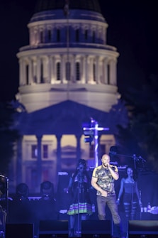 A singer performs energetically on a stage against the backdrop of an illuminated grand building. The stage is equipped with speakers and microphones, and two backup singers are visible. The building in the background is prominently lit, creating a striking visual contrast with the night sky.
