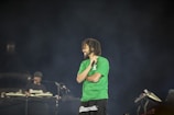 A man with a beard and short dreadlocks is holding a microphone on stage, wearing a green t-shirt, with another person in the background who appears to be operating DJ equipment. The background is dark, creating a spotlight effect on the man.
