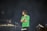 A man with a beard and short dreadlocks is holding a microphone on stage, wearing a green t-shirt, with another person in the background who appears to be operating DJ equipment. The background is dark, creating a spotlight effect on the man.
