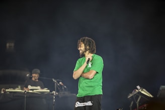 A man with a beard and short dreadlocks is holding a microphone on stage, wearing a green t-shirt, with another person in the background who appears to be operating DJ equipment. The background is dark, creating a spotlight effect on the man.