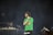 A man with a beard and short dreadlocks is holding a microphone on stage, wearing a green t-shirt, with another person in the background who appears to be operating DJ equipment. The background is dark, creating a spotlight effect on the man.