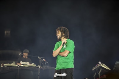 A man with a beard and short dreadlocks is holding a microphone on stage, wearing a green t-shirt, with another person in the background who appears to be operating DJ equipment. The background is dark, creating a spotlight effect on the man.