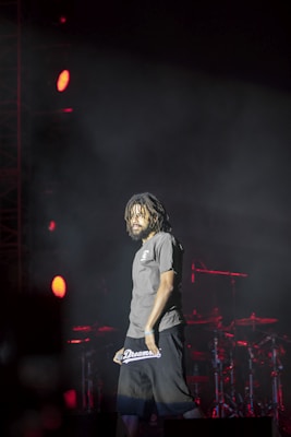 A man with dreadlocks stands on stage under dramatic lighting. He is wearing a gray shirt and black shorts with 'Dreamers' written on them. Red stage lights illuminate the background, highlighting the stage drum set and equipment.
