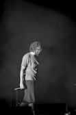 A person with shoulder-length dreadlocks stands on a stage, holding a microphone in hand. The image is in black and white, creating a dramatic and introspective atmosphere. Smoke or mist surrounds the person, enhancing the moody ambiance.