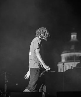A person with dreadlocks is on stage holding a microphone. The scene is dimly lit, suggesting a live performance or concert setting. The background includes part of a building structure, possibly a dome, partially illuminated.