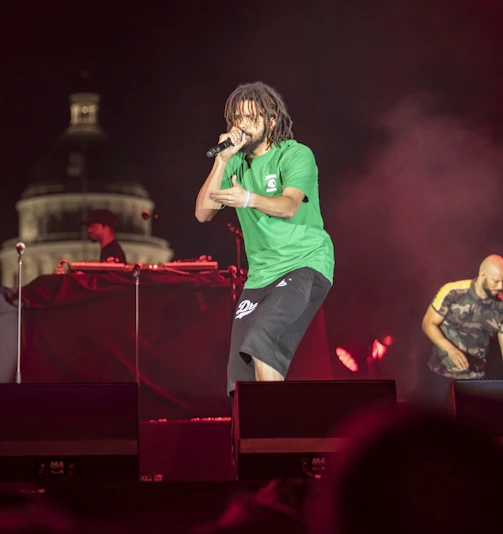 A dynamic shot of a rap artist performing passionately on stage with Buffalo city skyline glowing behind him at dusk.