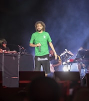 A male performer with dreadlocks wearing a green shirt and black shorts is on stage holding a microphone. The stage is set up with musical equipment, including keyboards and turntables. Three additional individuals are visible in the background, working with the equipment.