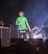 A male performer with dreadlocks wearing a green shirt and black shorts is on stage holding a microphone. The stage is set up with musical equipment, including keyboards and turntables. Three additional individuals are visible in the background, working with the equipment.