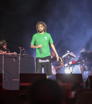 A male performer with dreadlocks wearing a green shirt and black shorts is on stage holding a microphone. The stage is set up with musical equipment, including keyboards and turntables. Three additional individuals are visible in the background, working with the equipment.