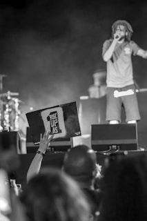 A person is performing on stage with a microphone in their hand, wearing casual clothing. In the foreground, an audience member is holding up a sign that reads 'RISE UP AS 1'. The background is blurred, showing parts of the stage and musical instruments.