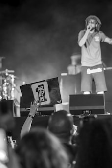 A person is performing on stage with a microphone in their hand, wearing casual clothing. In the foreground, an audience member is holding up a sign that reads 'RISE UP AS 1'. The background is blurred, showing parts of the stage and musical instruments.
