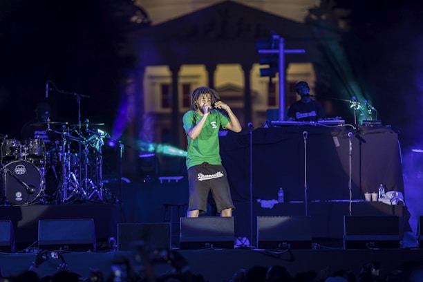 A concert scene features a performer in a green shirt and dark shorts energetically singing into a microphone. The stage is illuminated with colorful lights, and musical equipment including drums and a DJ setup are visible. In the background, a large building can be seen.